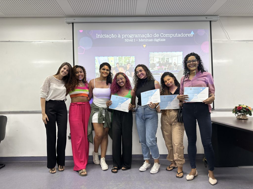 Sete mulheres jovens com aparências diversas e sorrindo, posam para foto. Quatro delas seguram seus certificados de conclusão do curso de Iniciação à Programação de Computadores, realizado na ACCS em parceria com o Programa Onda Digital.