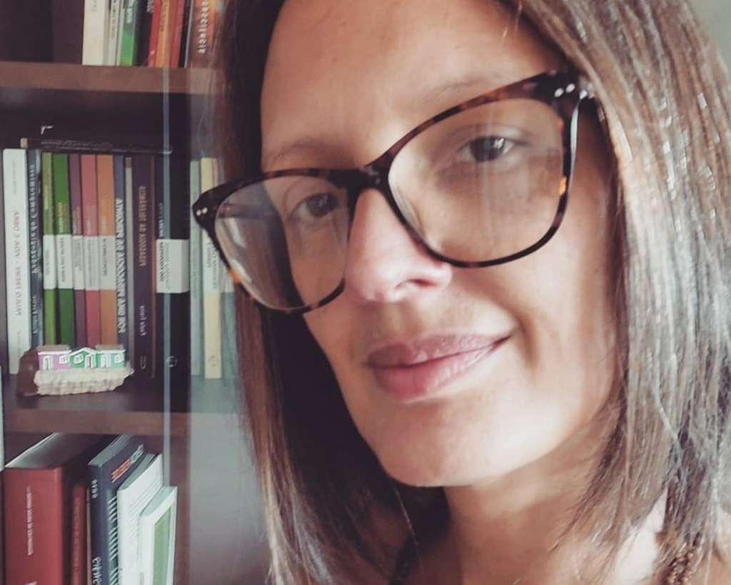 Mulher branca, de cabelos loiros-escuros. Usa óculos e esboça um leve sorriso no rosto. Ao fundo, estantes com livros.